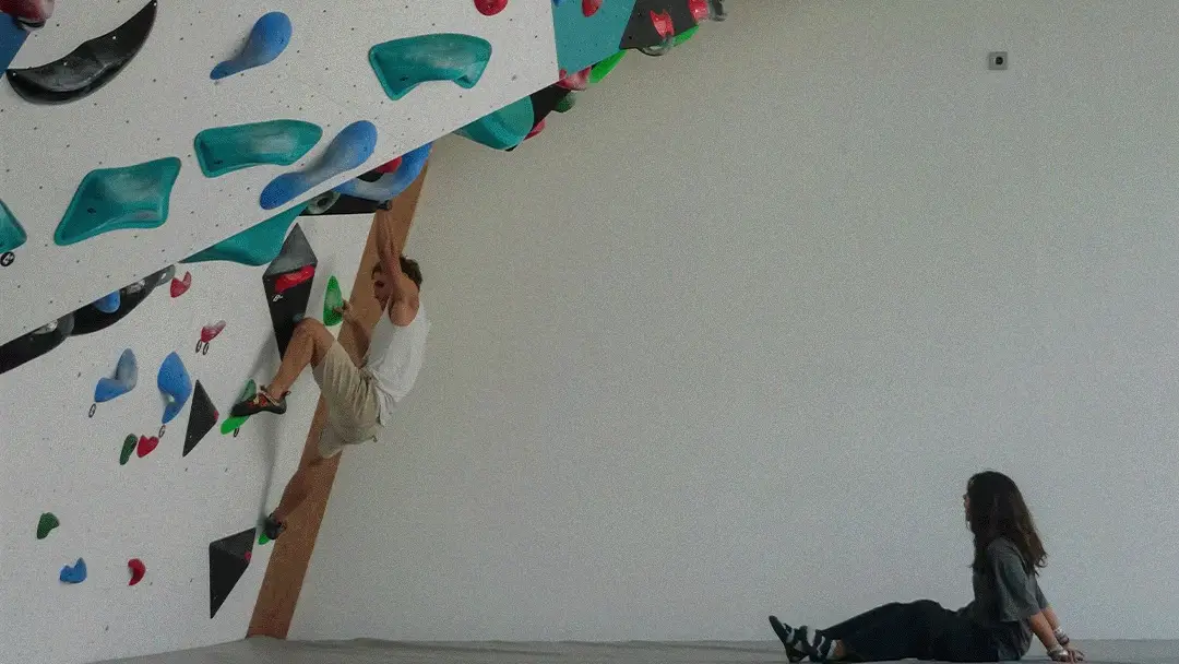 A climber hanging from holds on the overhanging bouldering wall at Bali Boulder in Sanur while a friend watches from the mat